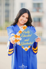 Graduate in a royal blue gown smiling while proudly displaying her decorated cap with floral details.