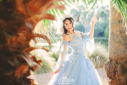 Dezeray smiling among palm branches, radiating joy in her pastel blue quinceañera dress during a photo session at Agua Caliente Park.