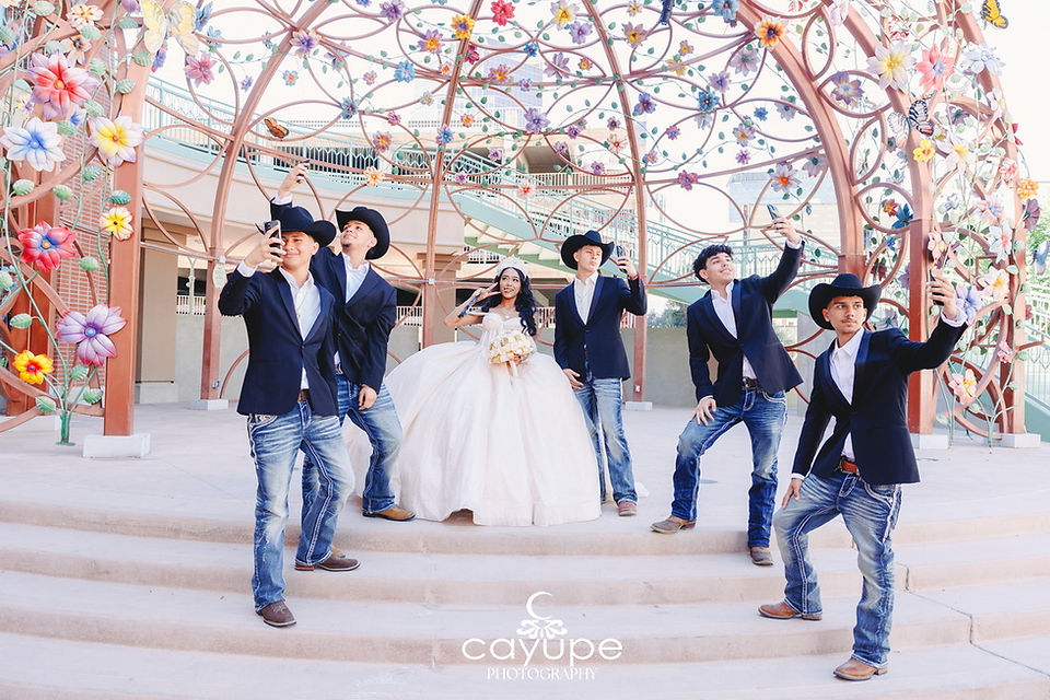Chambelanes wearing cowboy hats snap fun selfies with Ericka under a colorful flower canopy.