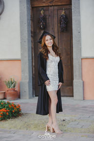 Brenda smiling, wearing a graduation cap and gown, standing in front of a rustic door.