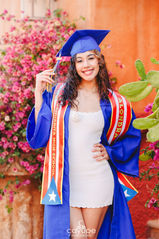 Cap and gown finale: Alondra smiles in full regalia, tassel in hand, against a warm terracotta wall and desert garden setting.