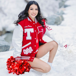 Jasmin kneeling in the snow, holding her pom-poms, wearing her cheerleading jacket, and smiling brightly.