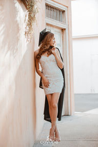 Brenda standing confidently, leaning on a rustic wall while wearing her graduation cap and a detailed white dress, with her gown draped over one arm.