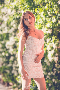 Brenda posing in a fitted white dress, surrounded by sunlit leaves.