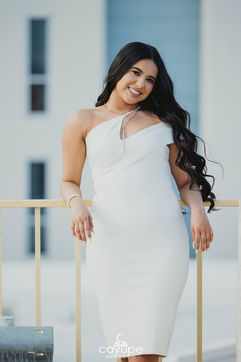 Close-up portrait of a young woman in a white dress leaning on a railing, smiling brightly with long wavy hair.