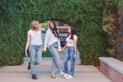 Diana and her two friends walk hand in hand under a green ivy-covered arch, smiling and enjoying the moment during her graduation photo shoot.