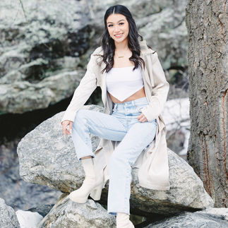 Jasmin seated on a large rock by a snowy forest, wearing a long beige coat and looking relaxed