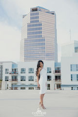 Elegant young woman in a white dress looking down while standing on a rooftop with a tall glass building behind her.