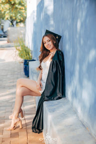 Brenda smiling radiantly while leaning against a blue wall, wearing her graduation cap and holding her gown over one shoulder.