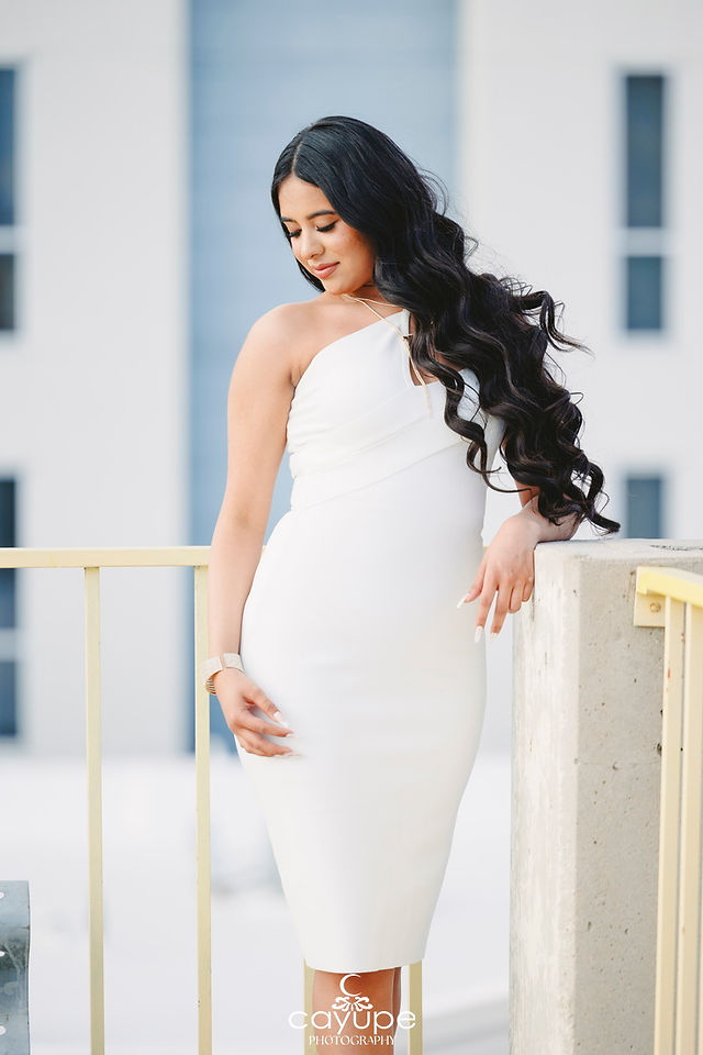 Young woman in a white dress looking down gracefully while leaning on a railing, with soft curls cascading down her shoulder.