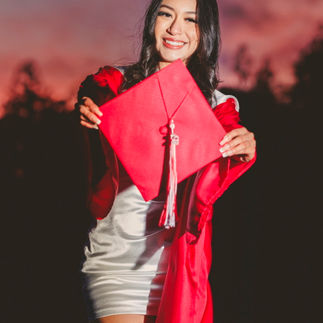 Jasmin holding her red graduation cap, smiling brightly with a stunning pink and purple sunset behind her.