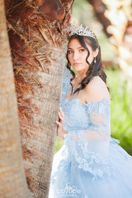 Dezeray looking at the camera beside a palm tree, wearing a delicate blue dress with lace details.