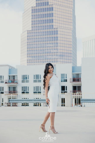 Young woman in a white dress standing on a rooftop, smiling softly with a cityscape in the background.