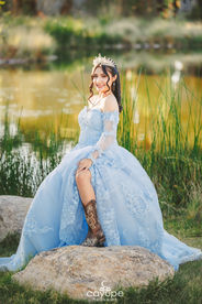 Dezeray smiling while sitting on a rock near the lake, pairing cowboy boots with her pastel blue dress.