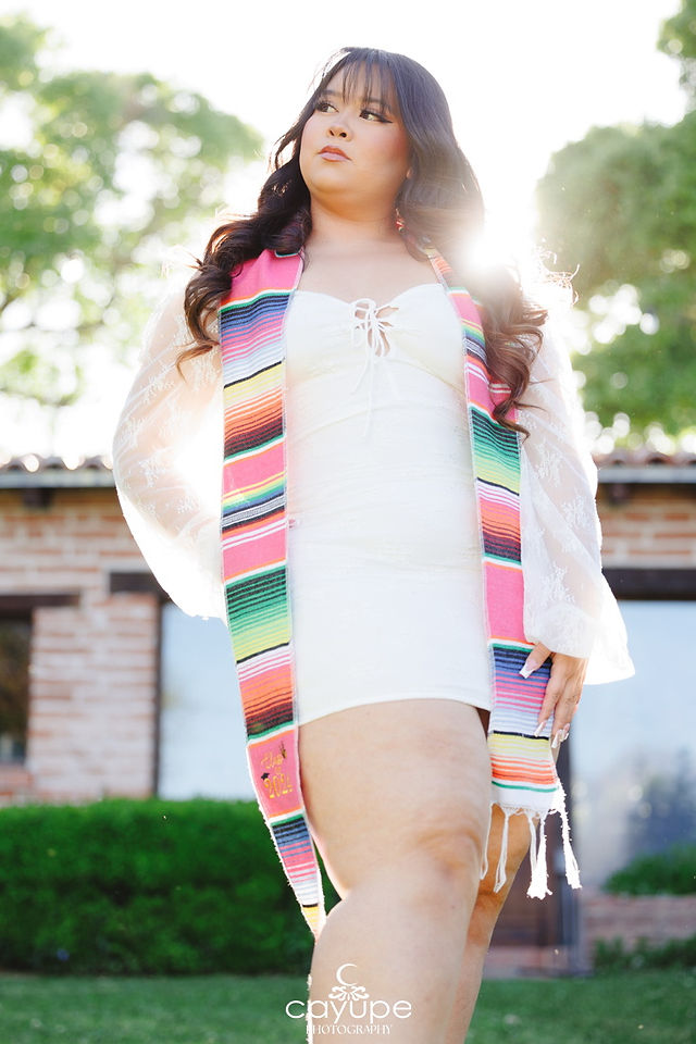 Empowering low-angle shot of Jasmine wearing a colorful serape stole and white dress, glowing with backlight