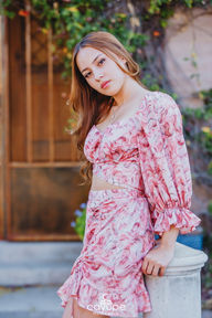 Brenda standing by a pillar, wearing a pink patterned outfit and showcasing a delicate necklace.