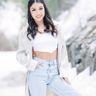 Jasmin wearing a long beige coat and jeans, standing in the snow with trees in the background, exuding a casual chic vibe.