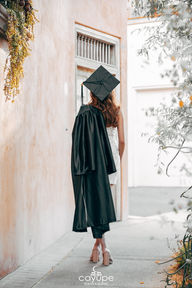 Brenda facing away from the camera in a graduation cap and gown, walking along a rustic pathway with soft greenery in the background.
