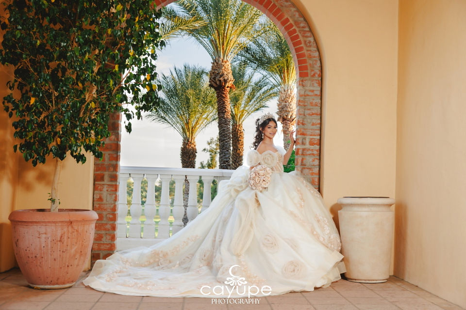Brianna stands framed by a brick archway, bouquet in hand, with a grove of palms and golden light behind her at Omni Tucson National Resort.