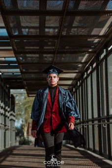 •	Wearing a navy graduation gown and cap over his red velvet blazer, Christian strides across a metal pedestrian bridge, marking his senior milestone.