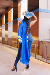 Graduate in a black top and jeans holding her decorated cap up high, draping her blue gown over her shoulder, with a modern building in the background.