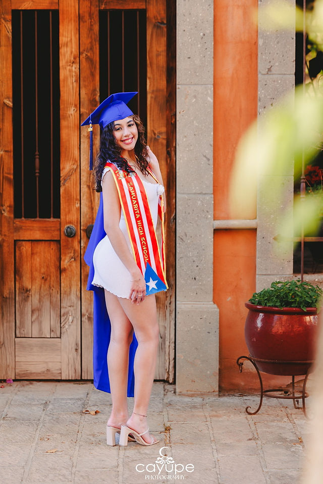 Alondra stands in full Sahuarita High School regalia—royal blue cap, flowing gown, and red and yellow stole—smiling outside rustic wooden doors.