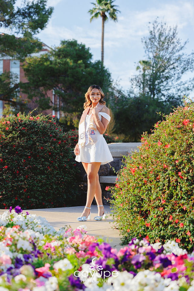 Cassandra standing in a white dress with a graduation stole in a garden of colorful flowers at the University of Arizona.