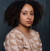 A portrait of a multi racial woman with curly hair, wearing a beige and wheat coloured hounds tooth styled crew neck sweater. The background is dark blue.