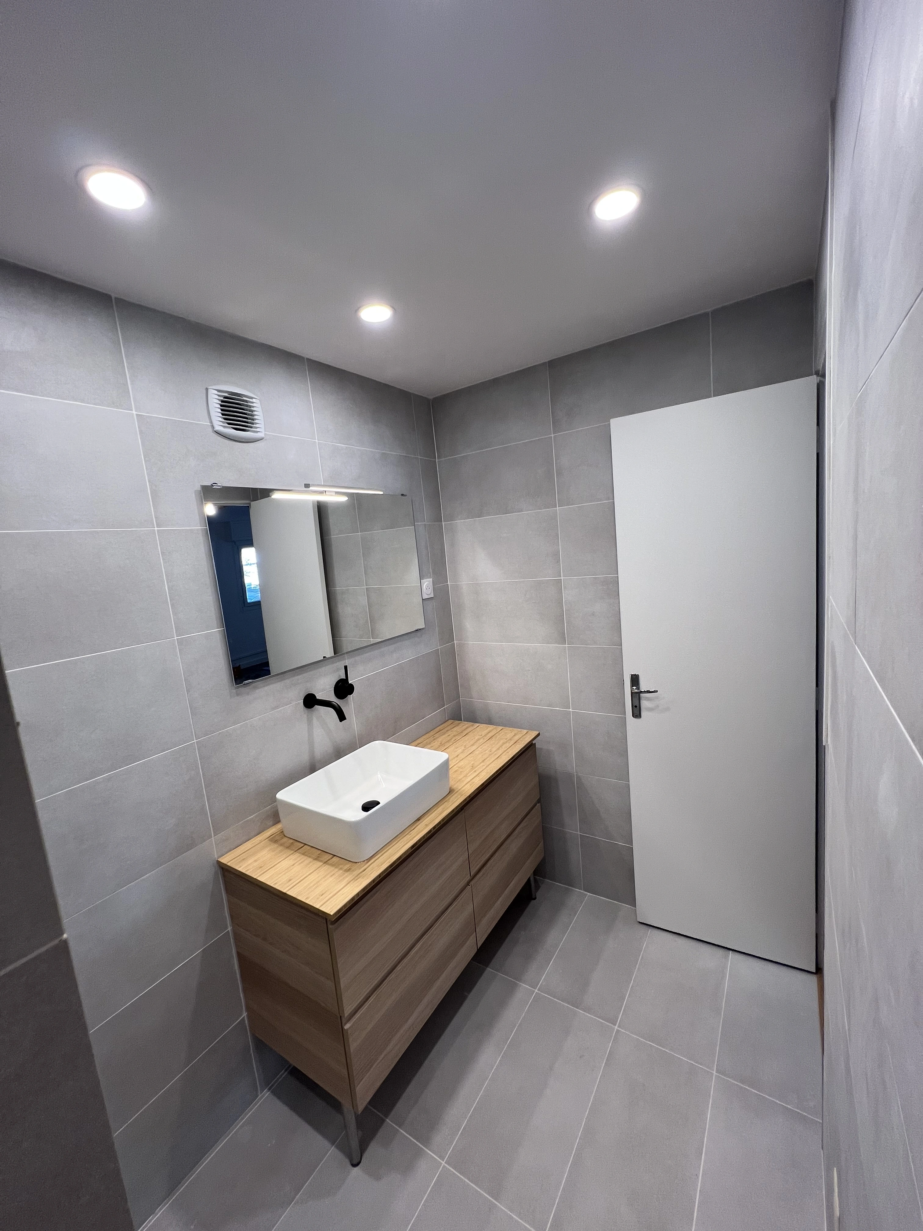 Vue rapprochée d'une salle de bain moderne avec carrelage gris, meuble vasque suspendu en bois clair, vasque blanche rectangulaire et robinetterie noire.  Design épuré et fonctionnel.