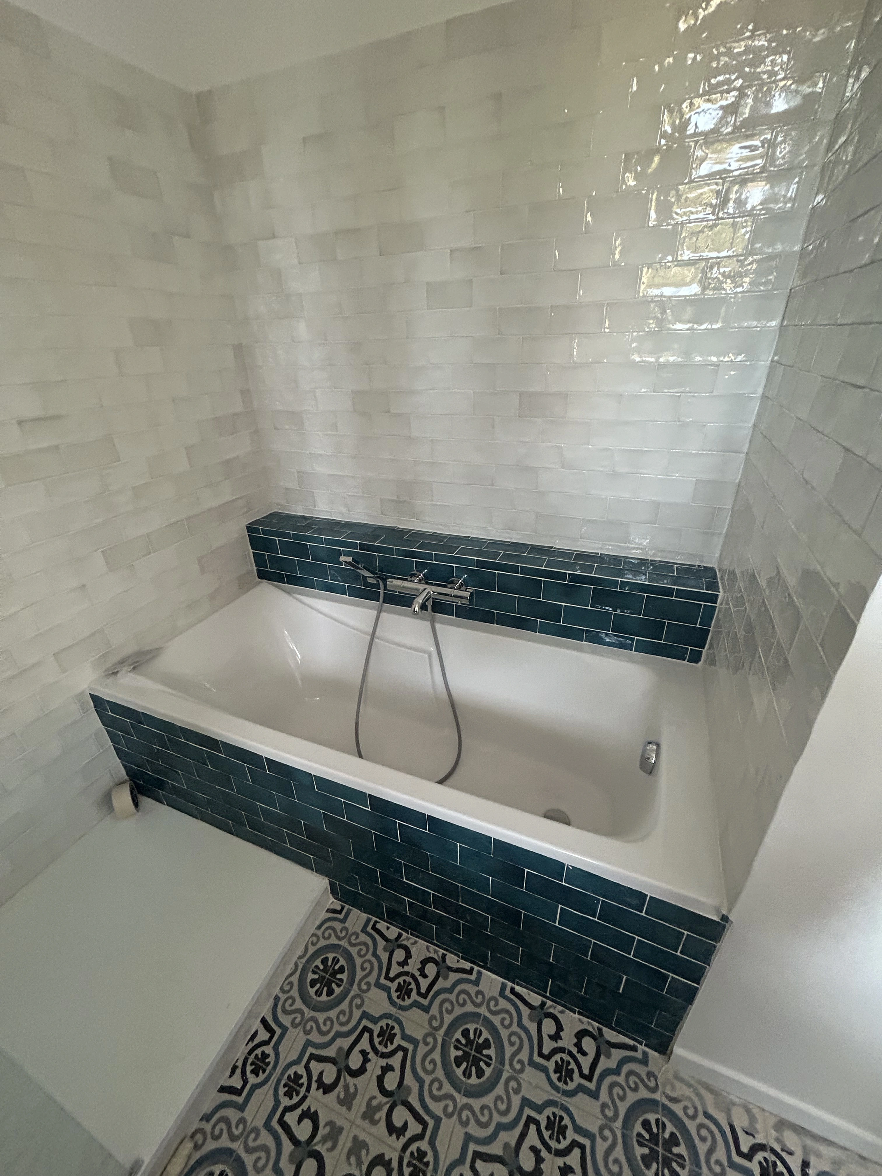Salle de bain avec murs en carrelage imitation zellige blanc, sol en carrelage à motifs bleus et blancs, et baignoire blanche.  Focus sur la texture des carreaux et les joints.