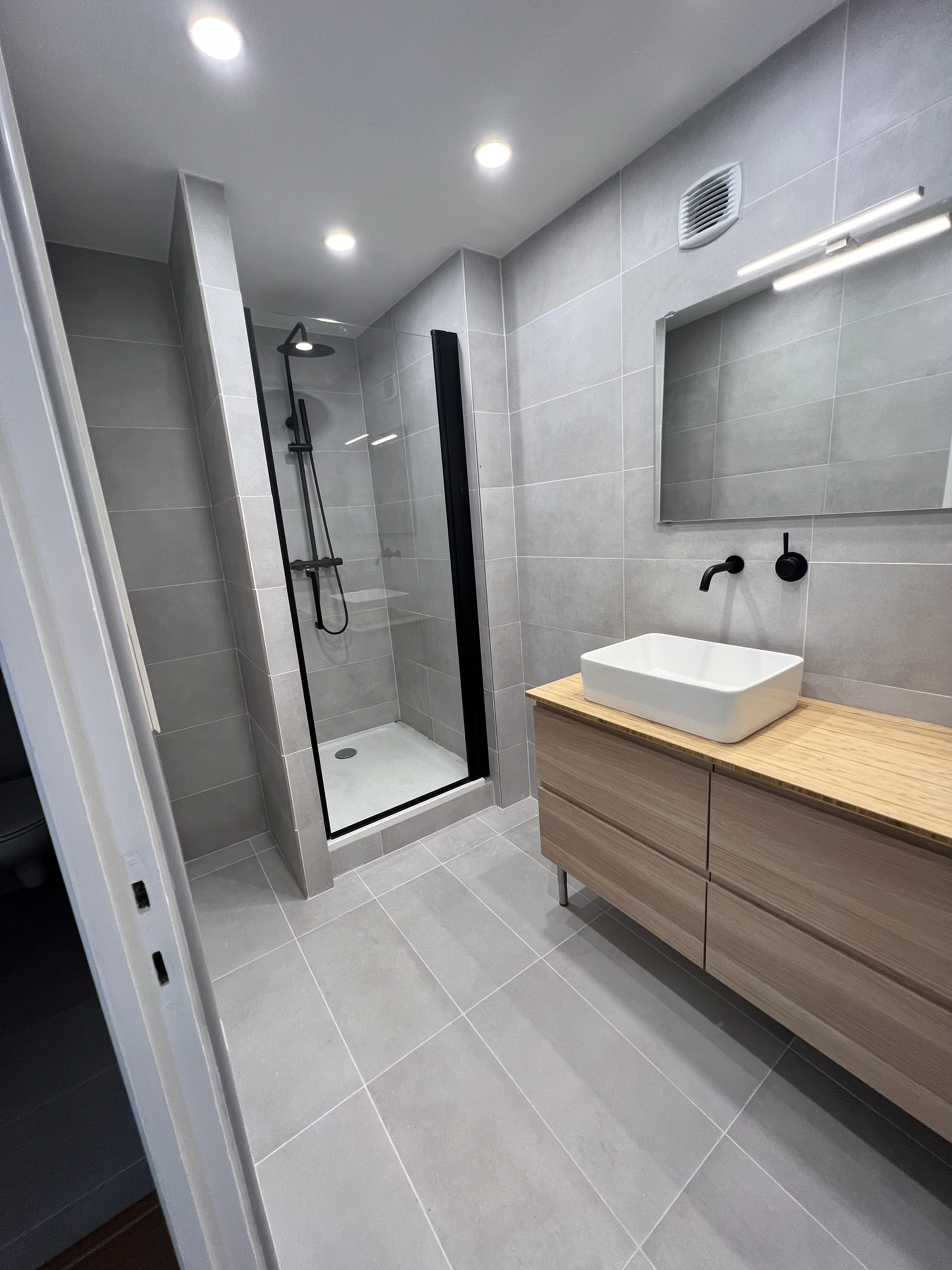 Salle de bain contemporaine rénover avec carrelage gris, douche spacieuse avec paroi en verre, meuble vasque en bois clair avec vasque blanche et robinetterie noire.  Design moderne et élégant.
