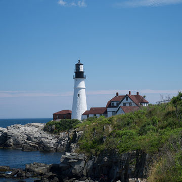 A white lighthouse stands on rocky cliffs by the ocean, with red-roofed buildings and greenery around it under a clear blue sky.