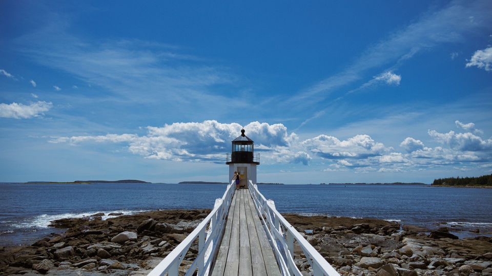 A lighthouse at the end of a wooden walkway, with a clear blue sky and calm ocean in the background, surrounded by rocky shores.