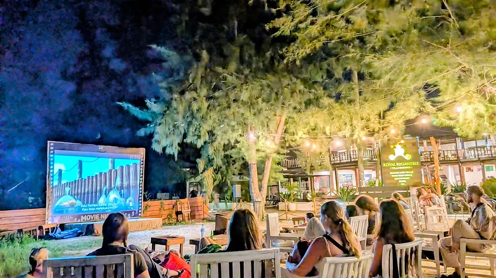 People watch an outdoor Cinema on the beach with a lit resort patio under trees, with a screen, string lights, at Royal Regantris Gili Air