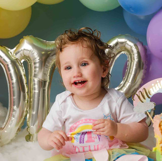 cakesmash with baby smiling in front of rainbow coloured balloons.