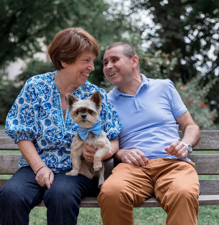 photographie d'un petit chien avec un couple de personne âgé assis sur un banc dans un parc à la rochelle. le couple se regarde et rigole. le petit chien est au milieu avec un nœud papillon bleu 