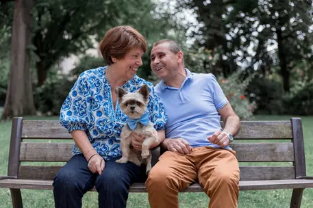 photographie d'un petit chien avec un couple de personne âgé assis sur un banc dans un parc à la rochelle. le couple se regarde et rigole. le petit chien est au milieu avec un nœud papillon bleu