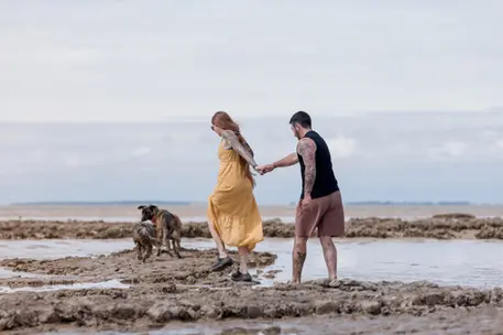 photo d'un couple qui se tient la main, la femme est devant et tire l'homme du bras pour le guider. ils marchent sur la plage pleine de boue avec 2 chiens devant eux