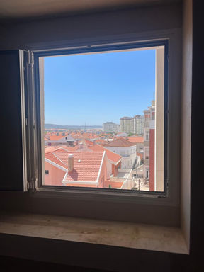 View through a hotel room window overlooking rooftops and the Costa da Caparica coastline.