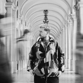 A person with fair skin stands confidently in the center of a bustling hallway, surrounded by a flurry of activity as people move around them.