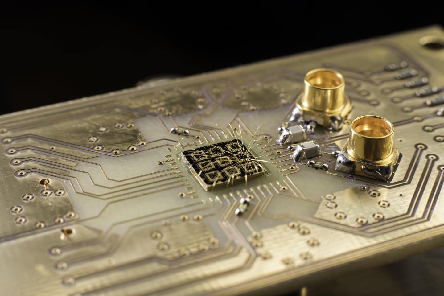 Thin aluminum wires connect the surface of a quantum processor semiconductor chip to pads on a circuit board. The researchers fabricate the device by patterning and depositing metal gates on a chip. The metal gates are designed to trap individual electrons in the semiconductor. The researchers send electrical signals to the device via the aluminum wires, changing the voltage on the metal gates to control the electrons. They also receive electrical signals from the device to help monitor the electrons’ behavior.@ University of Rochester / J. Adam Fenster