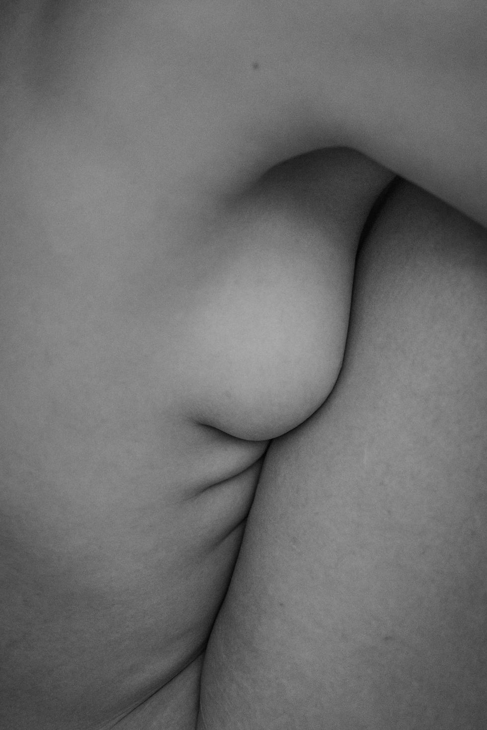 Black and white photo of nude female body with only the mid section showing from the side with legs folded up toward chest covering the breast and revealing un-retouched fat folds and a few stretchmarks