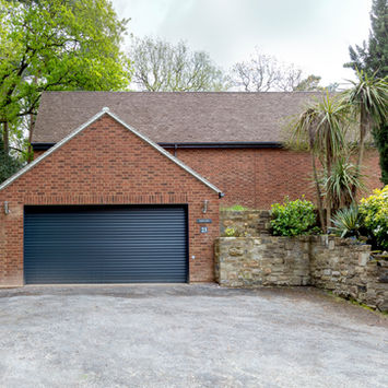 Brick garage with closed dark door and driveway, surrounded by greenery.