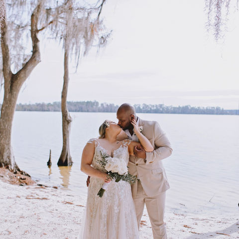 Newlywed couple embracing on sandy shore by serene lake with mossy trees.