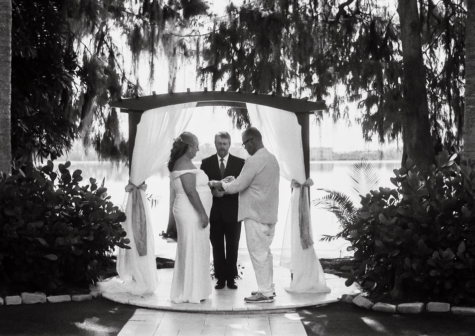 Black and white wedding ceremony by a lake with bride, groom, officiant.