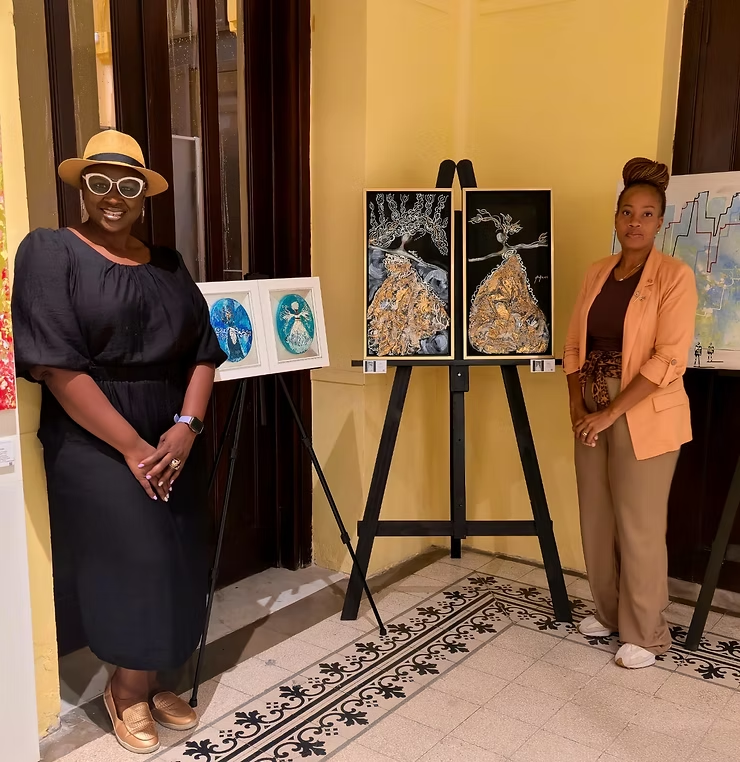 Two women in a yellow-walled gallery stand beside easels displaying blue-toned and gold dress-themed abstract artworks.