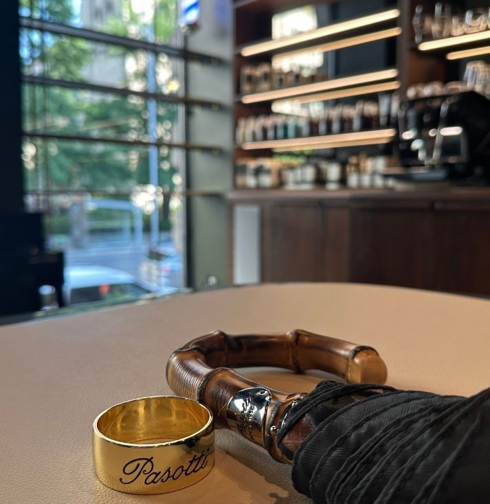 Handmade Pasotti Italian umbrella with bamboo handle and gold-finished ring displayed on a genuine leather presentation island at ÜLRIC men's grooming atelier in Montréal, with the Sanremo espresso bar and curated retail collection in the background.