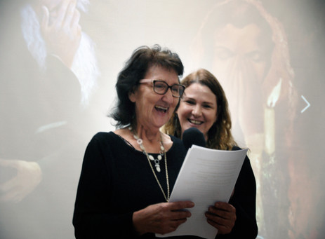 Two women smiling, one speaks into microphone at Elkana exhibition.