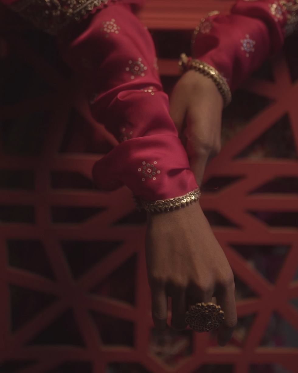 A close-up image of a person's hands wearing a pink, embroidered sleeve with gold jewelry, including a gold bracelet and a large, ornate gold ring. The background features a geometric pattern in a reddish hue.