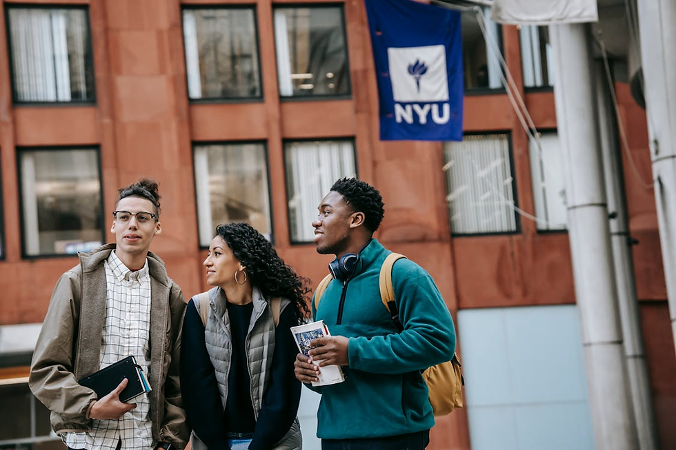 Three people stand smiling outside a red building with an NYU flag. They hold books and wear casual jackets, evoking a lively college scene.